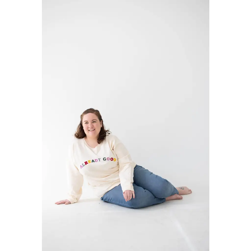 Person in cream embroidered organic cotton sweatshirt and blue jeans sitting on floor