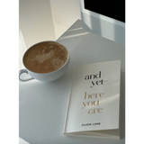 Cozy white cup of hot chocolate with Eileen Lamb’s And Yet Here You Are book, Edwardsville