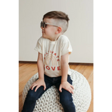 Child in sunglasses and LOVE shirt on a pouf, showcasing Cultivate Love Organic Cotton