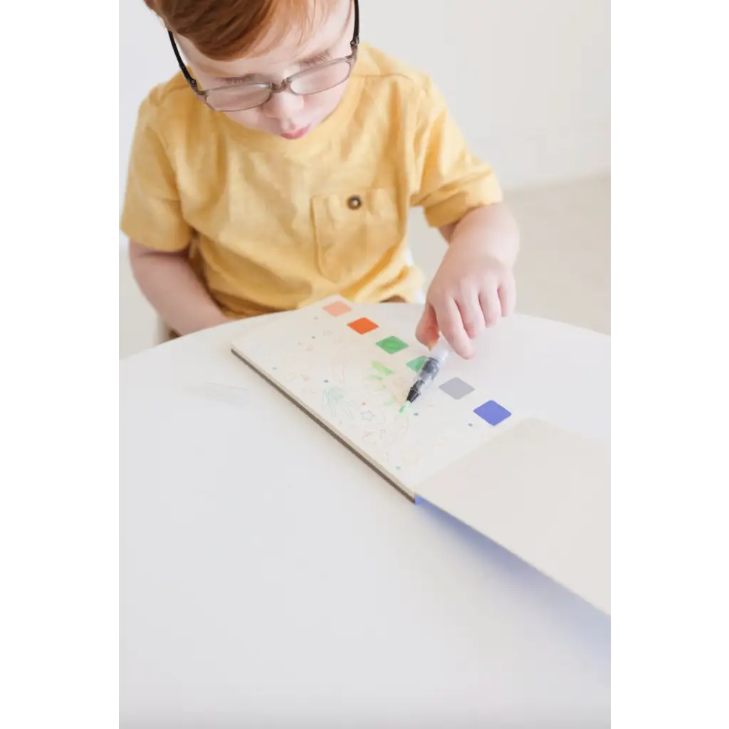 Young kid in yellow shirt with watercolor book and colorful prints, holding a brush pen