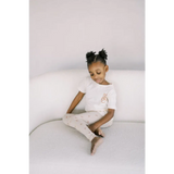 Child in white t-shirt and light pants sits cross-legged in Ditsy Rainbow Leggings set