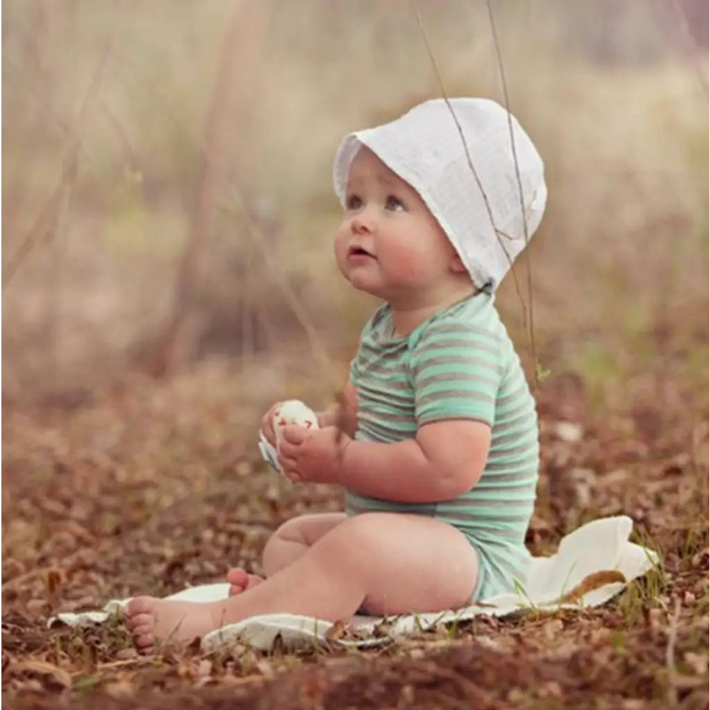 Baby in a white bonnet and green striped onesie with Estella Organic Baseball Rattle