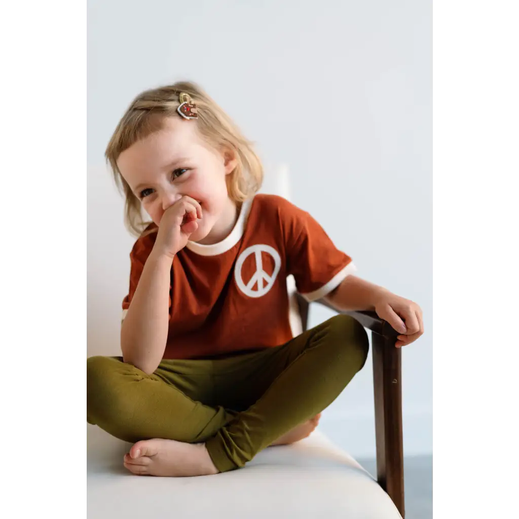 Giggling kid in rust Peace Embroidered Ringer Tee and olive pants, perfect for everyday fun