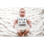 Baby in a white onesie saying TALK OF THE TOWN, made of soft organic cotton