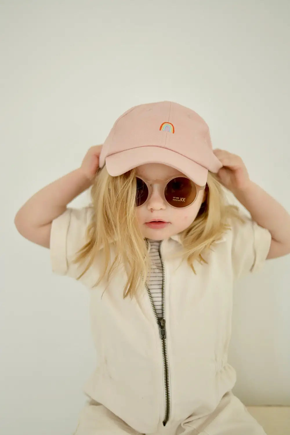 Kid in a pink baseball cap and round sunglasses rocking a cool Henley shorty romper.