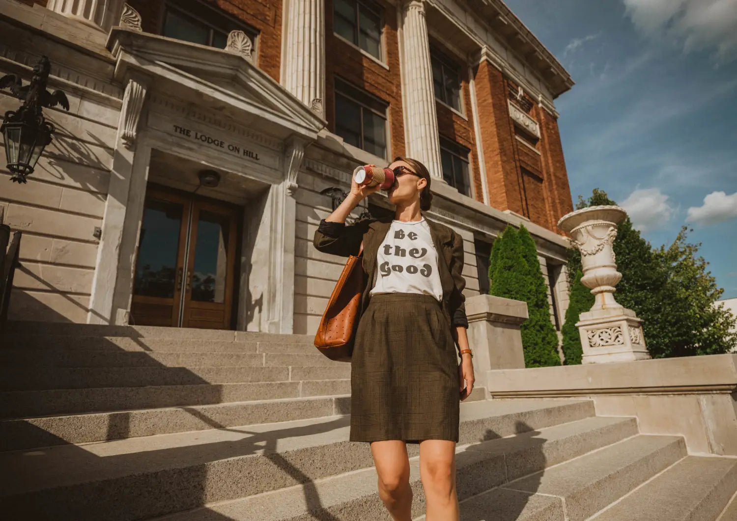 Person in a brown jacket and dark skirt enjoying a drink on stone steps, stylish vibe for the grind worth.