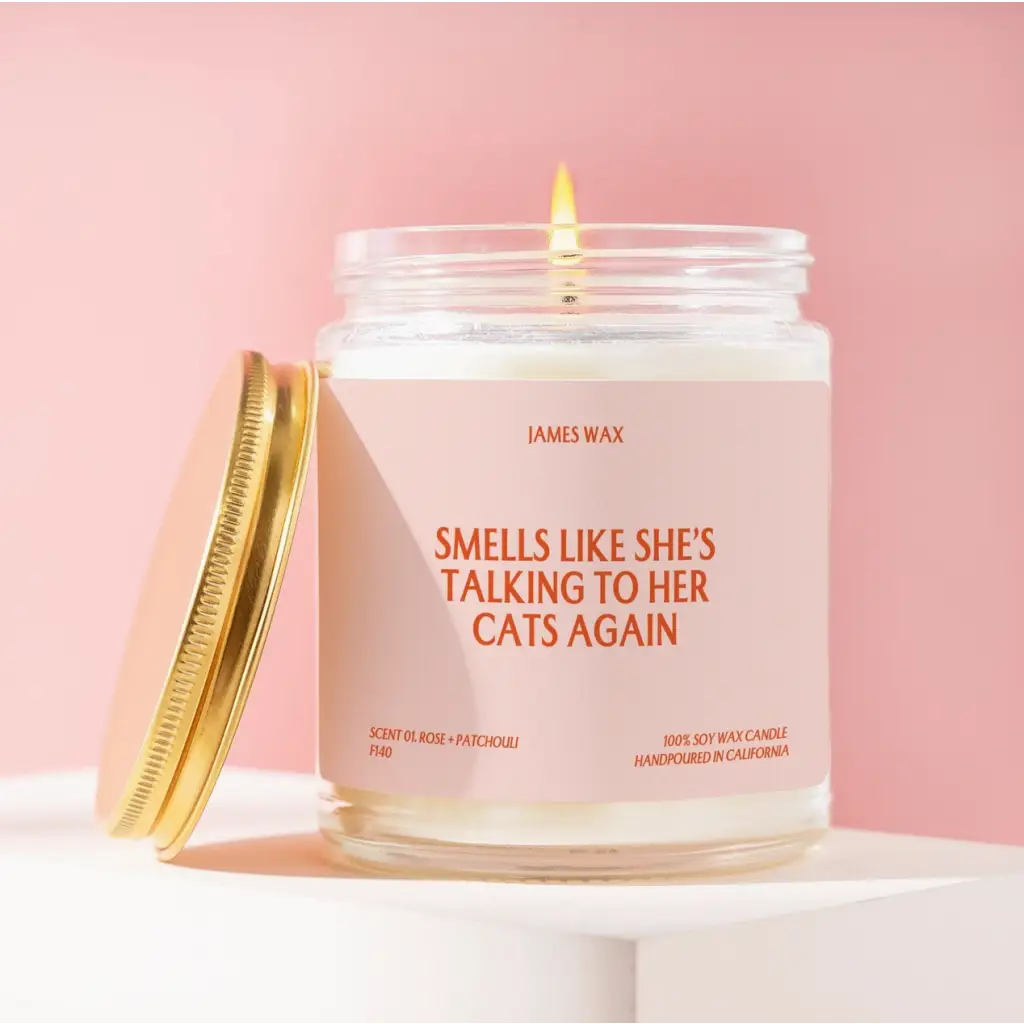 White candle in a jar with gold lid, featuring humorous cat talk text, Rose + Patchouli scent