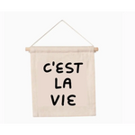 Cream banner with black text saying CEST LA VIE on natural canvas with wooden rod hang sign