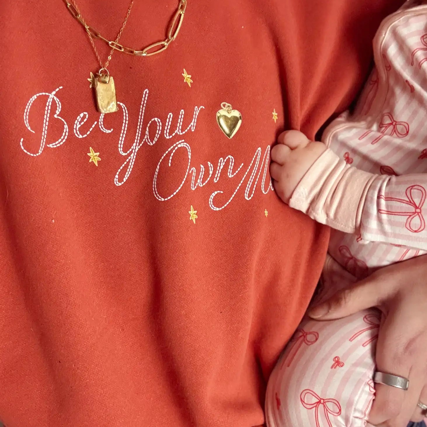 A coral-colored sweatshirt with ’be your own’ embroidered in white cursive script, accented by gold star and heart charms.