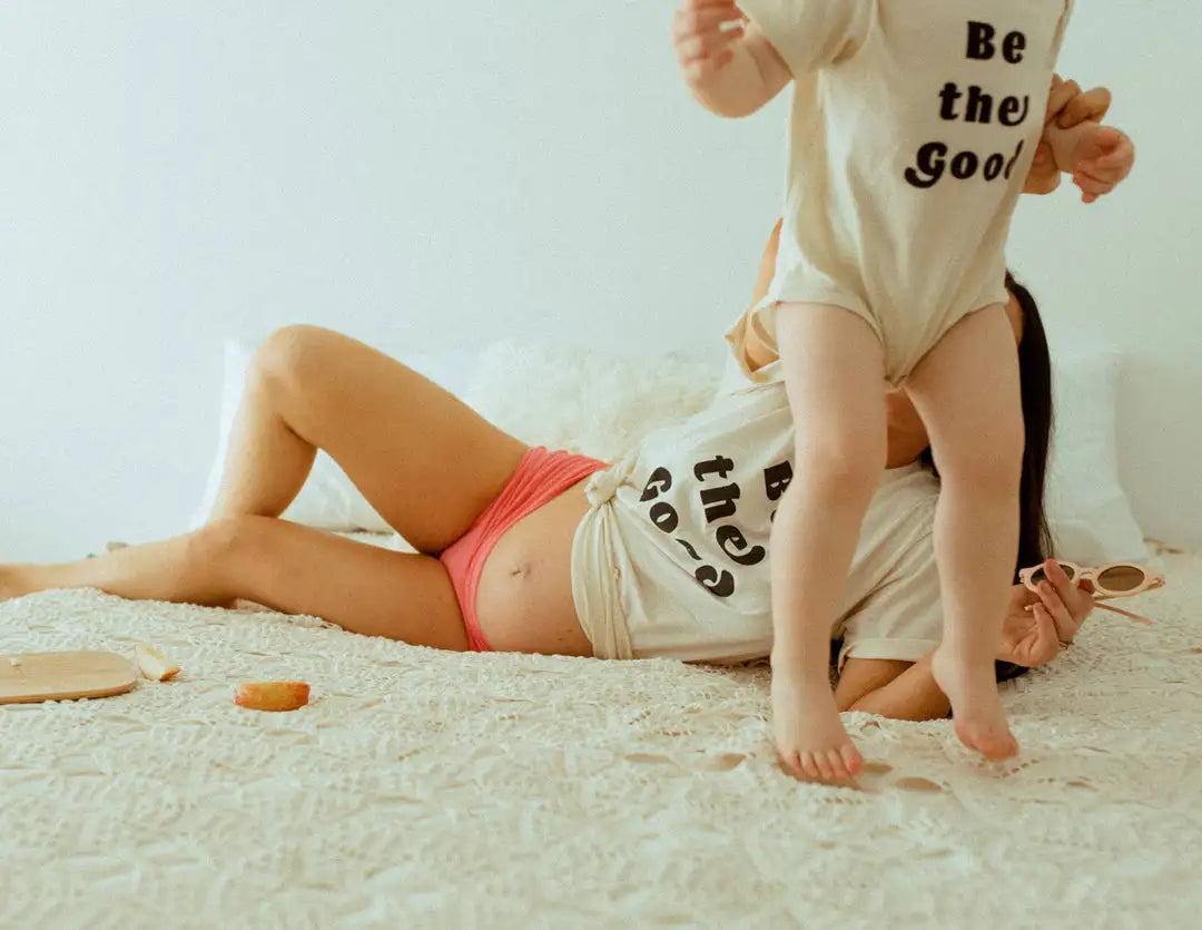 A cream-colored, short-sleeved baby onesie with black printed text reading ’be the good.’
