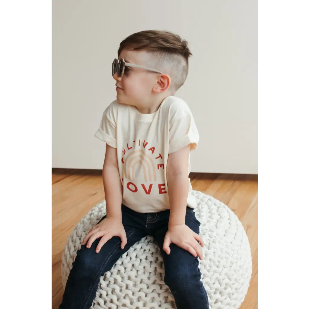 Child in sunglasses and LOVE shirt on a pouf, showcasing Cultivate Love Organic Cotton