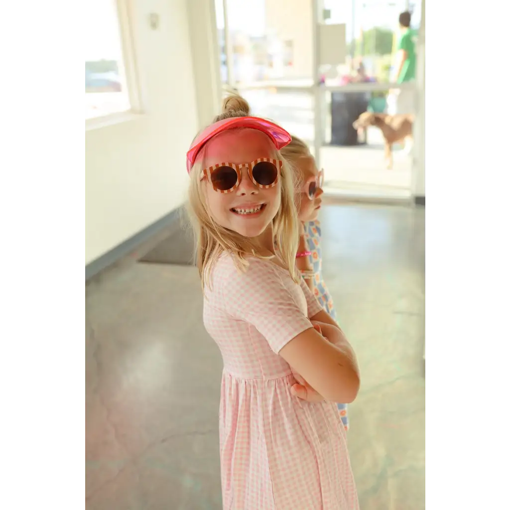 A happy child in a pink Gingham All Day Twirl Dress, red cap, and heart sunglasses