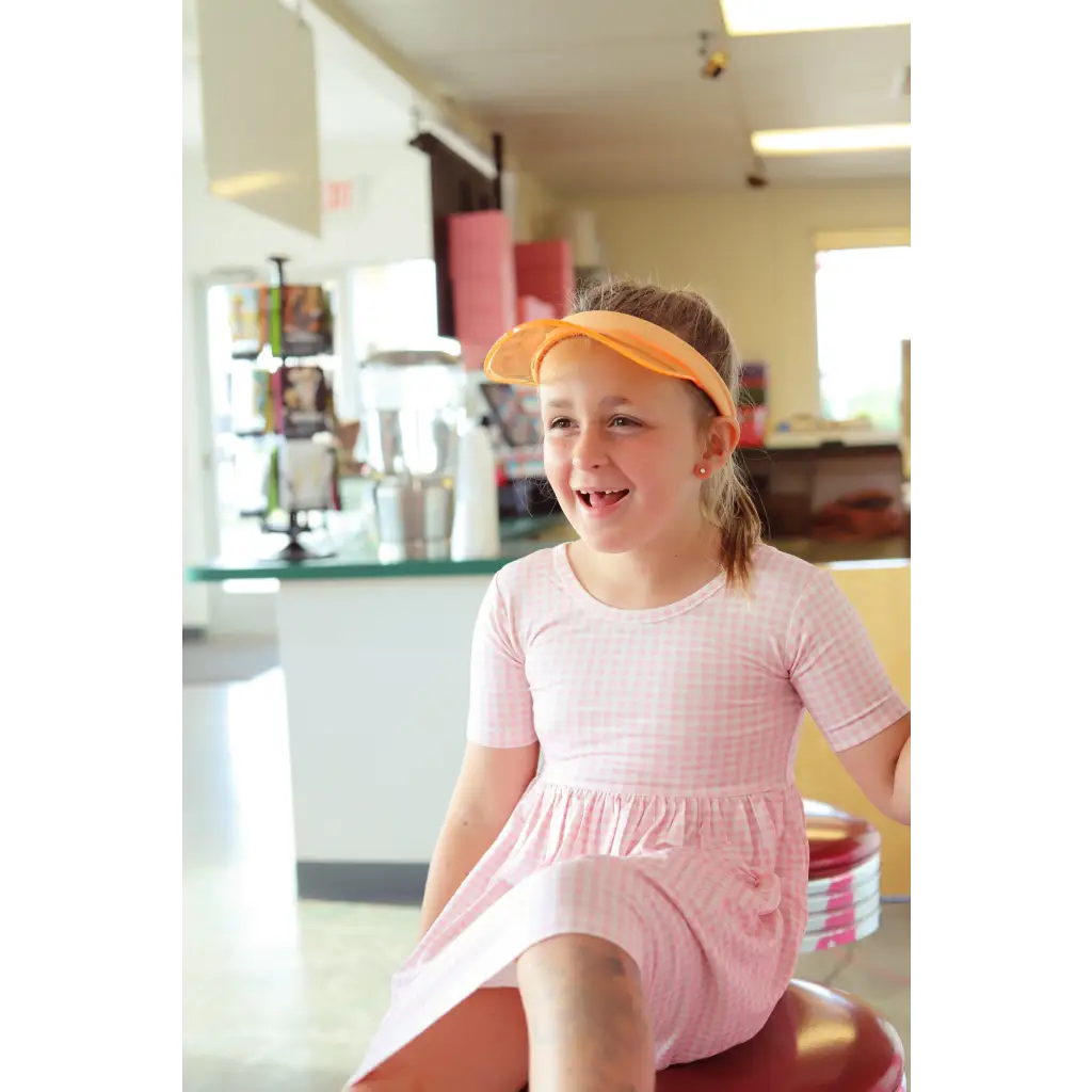 Child in a pink twirl dress and orange visor sits on a stool for Gingham All Day Twirl