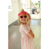 Child in pink gingham dress sporting a red cap and heart sunglasses, stylishly fun!