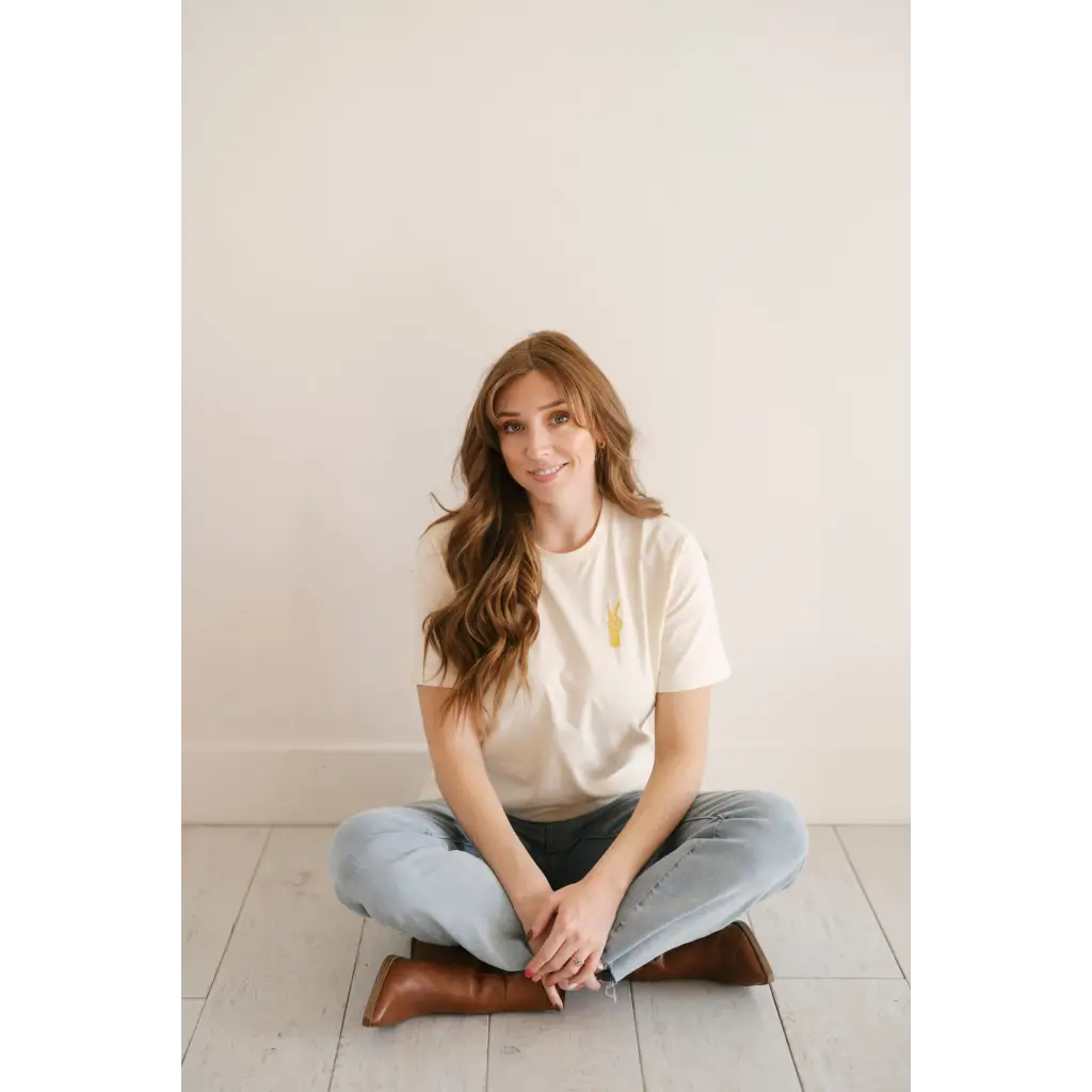 Person in light blue jeans and White Good Vibes Peace Embroidered Tee sitting cross-legged