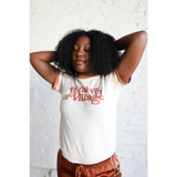 Person in a white graphic Adult Retro Ringer Tee and rust pants, rocking curly hair