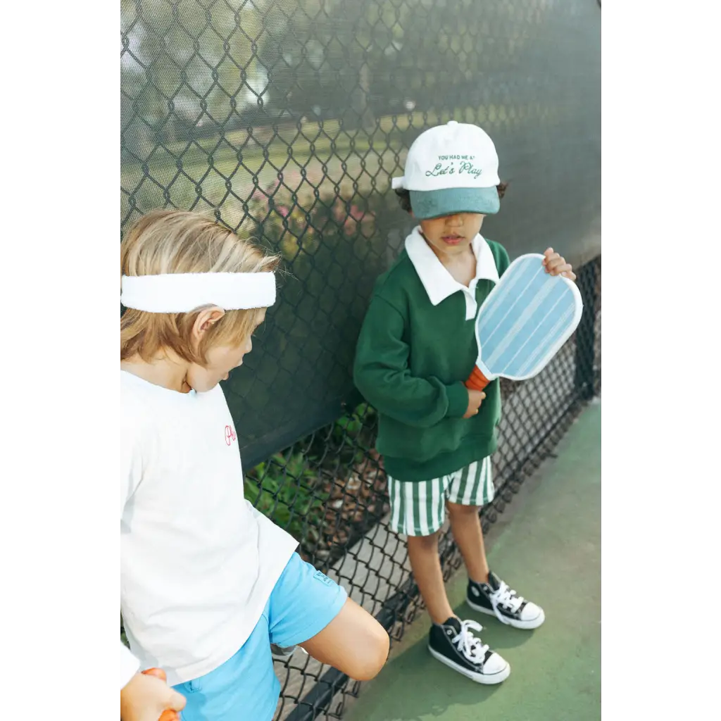 Two kids in sneakers by a fence showing off their mini pickle ball paddles