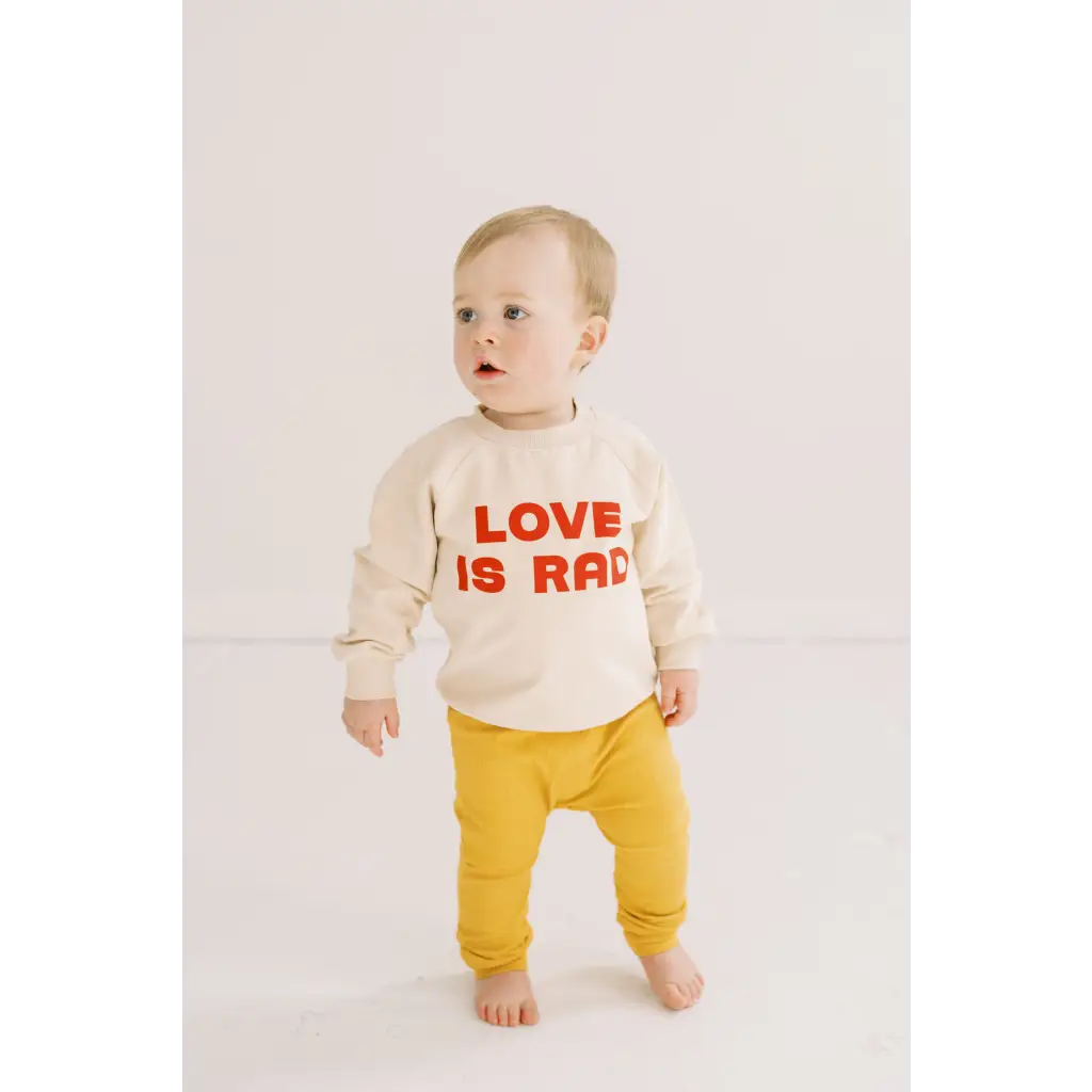 Toddler in cream Love Is Rad baby pullover sweatshirt and yellow pants, made of organic cotton fabric