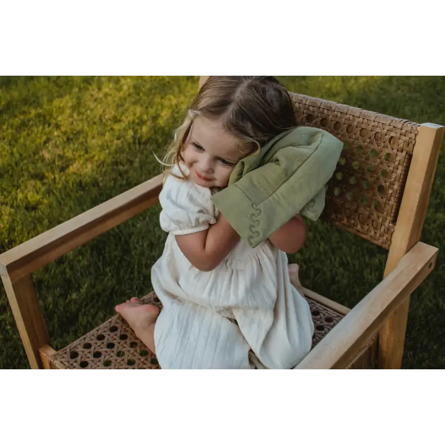 Child in a white dress on a wooden chair with a Mini Cozy Organic Cotton blanket