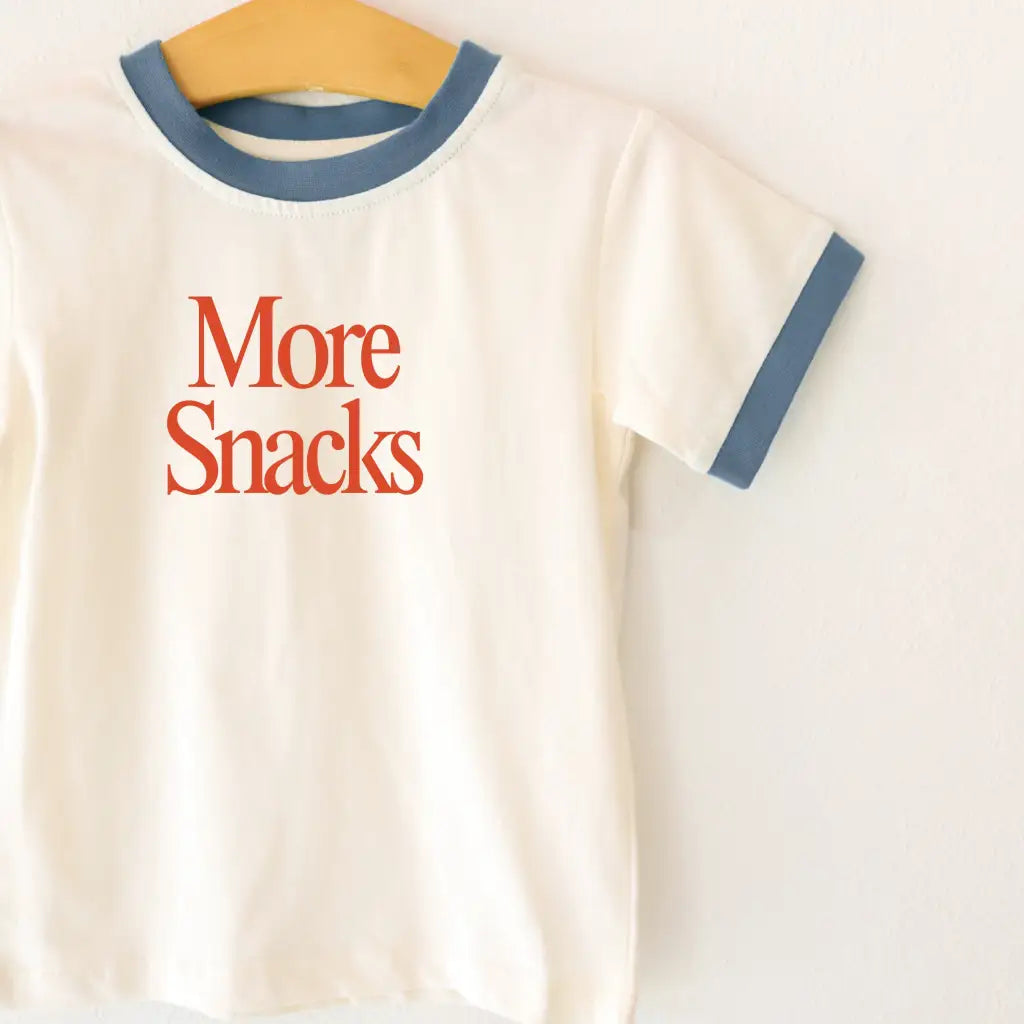 Cream-colored kids ringer tee with navy trim and bold orange More Snacks text