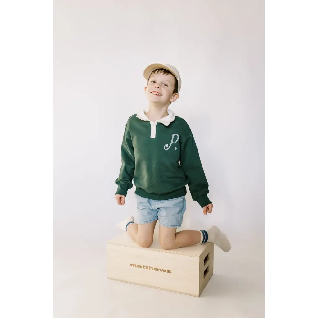 Child in a green kids embroidered polo pullover, kneeling on a wooden box