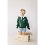 Child in a green kids embroidered polo pullover, kneeling on a wooden box