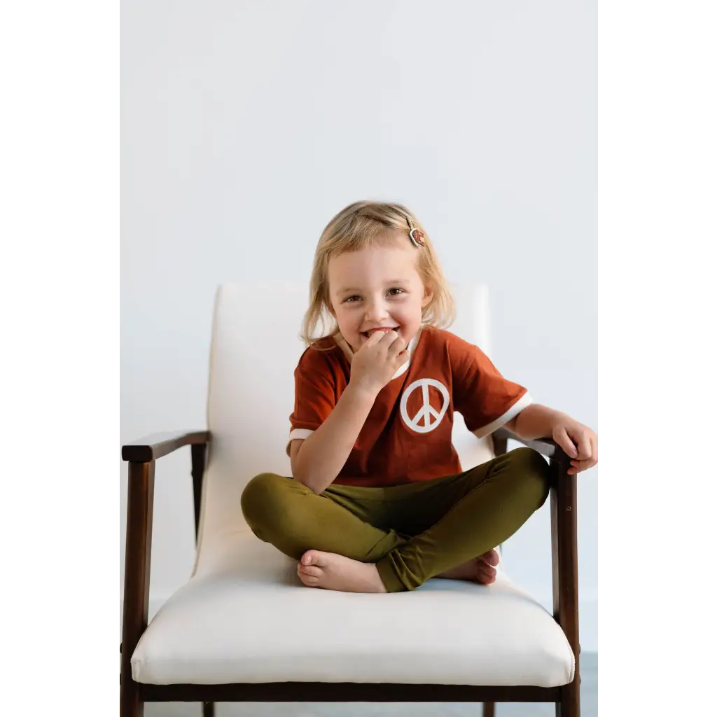 White upholstered chair with wooden armrests next to a kids Peace Embroidered Ringer Tee