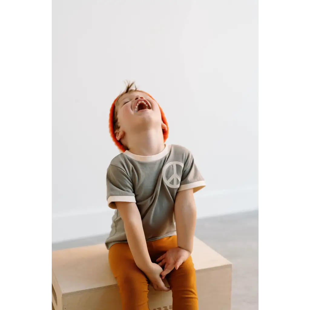 Laughing child in gray peace embroidered ringer tee and orange pants, ready to play
