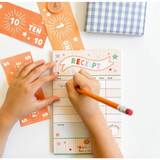 Hands writing on a pretend play receipt with orange number cards for creative kids