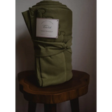 Folded olive-green organic cotton blanket on a wooden stool with a label