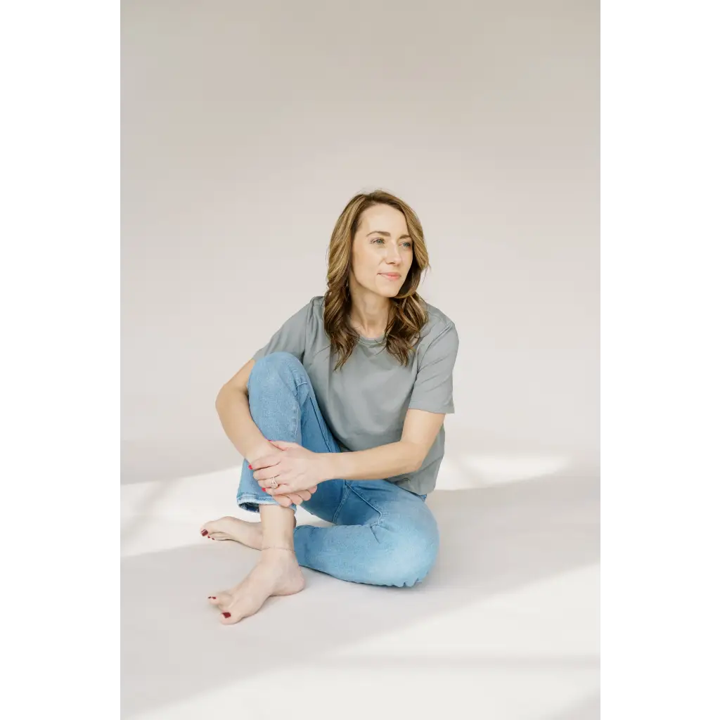 Person in a grey mom crop boxy tee and blue jeans sitting cross-legged on the floor