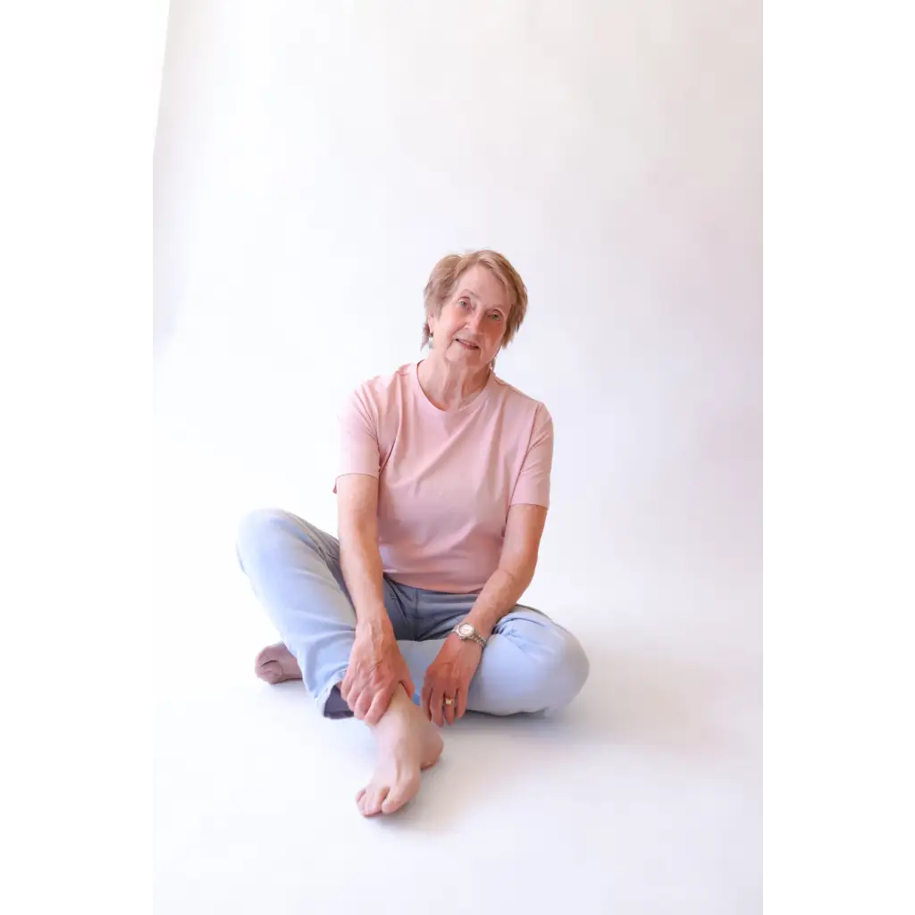 Older woman in pink shirt and jeans sitting cross-legged in The Mom Crop with polished prints