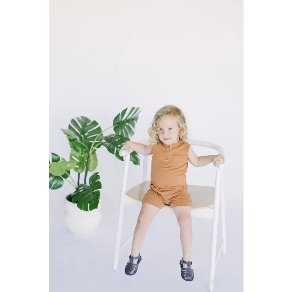 Cute child in brown wearing a Henley Shorty Romper on a white wooden chair