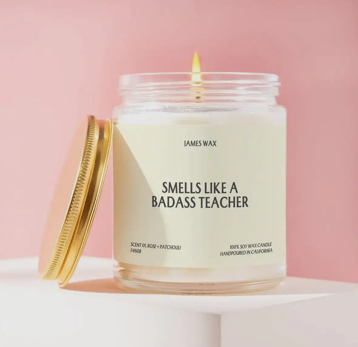 White scented candle in a glass jar with a gold lid and humorous ’Smells Like A Badass Teacher’ label.