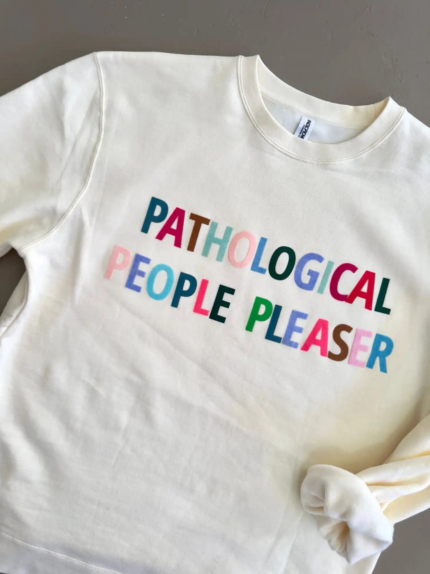 White sweatshirt with colorful text reading ’PATHOLOGICAL PEOPLE PLEASER’