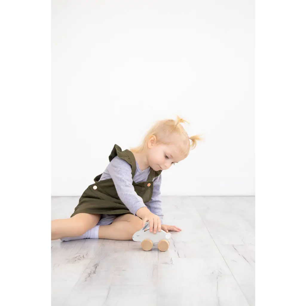 Cute olive green kids overall dress with white trim beside wooden bunny push toy
