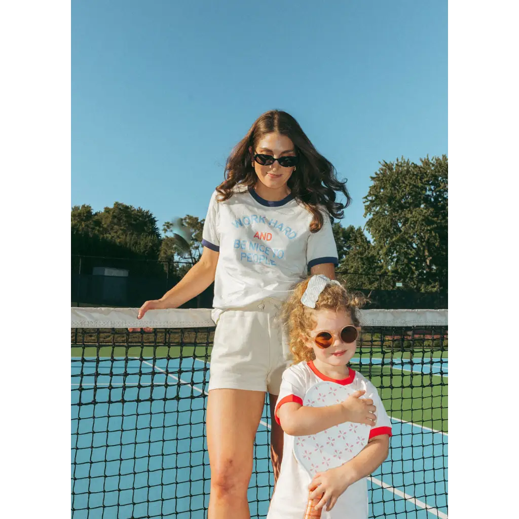 Two people in matching white athletic outfits by a tennis net wearing Word Hard Unisex Ringer